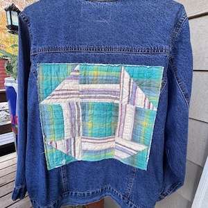 May include: A dark blue denim jacket featuring a large, square, quilted fabric patch sewn onto the back. The patch displays a geometric pattern in shades of teal, white, and purple plaid, with visible stitching throughout the quilted fabric surface.