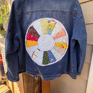 May include: A blue denim jacket featuring a large, circular, multi-colored patchwork applique on the back. The circular design consists of various fabric segments in floral, plaid, and solid patterns, surrounding a plain white center circle.