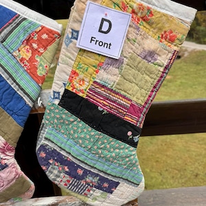May include: Two patchwork Christmas stockings with a variety of fabric patterns and colors, including floral, striped, and solid designs. One stocking has a tag that reads "D Front". The stockings are displayed on a wooden surface.