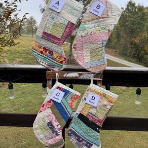 May include: Four handmade Christmas stockings with patchwork designs. Each stocking features a unique combination of fabrics in various colours, including pink, green, blue, and white. Each stocking has a white tag that says "Front".