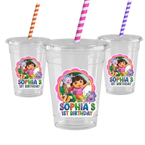 May include: Three clear plastic cups with lids and straws. Each cup has a colorful sticker featuring Dora the Explorer and the text "Sophia's 1st Birthday." The straws are orange, pink, and purple with white stripes.