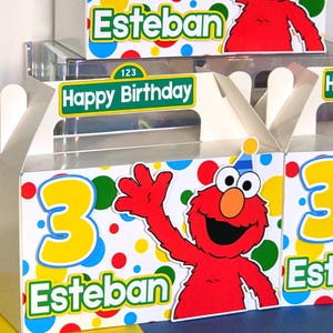 May include: Birthday party favor boxes featuring the character Elmo. The boxes are white with colorful polka dots, a large yellow number 3, and the name "Esteban". The boxes also have a "Happy Birthday" sign.