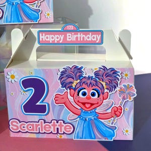 May include: A white party favor box with a cartoon character illustration, the number 2, and the name "Scarlette". The box has a "Happy Birthday" sign and a handle. The background is pink with a rainbow design.