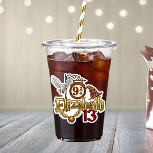 May include: A clear plastic cold cup filled with dark liquid and ice, featuring a white and gold striped straw. The cup displays a decal with the name Elizabeth, the number 13, a white owl, a brown sorting hat, a golden snitch, and a Platform 9 3/4 sign.