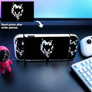Op de afbeelding: Een zwart-witte Nintendo Switch-console met een grafisch ontwerp. Het ontwerp toont een witte illustratie op een zwarte achtergrond. De console staat op een donkere ondergrond met een toetsenbord en controller zichtbaar.