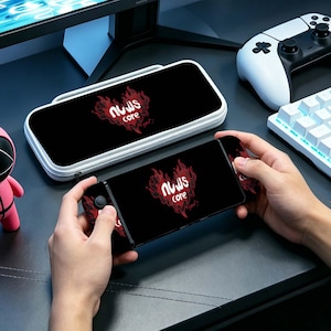 Peut inclure: Console de jeu noire avec un logo rouge et blanc, tenue en mains. Une housse de transport assortie est également visible. Le logo présente un motif en forme de cœur avec des flammes et le texte "nuws core". Un clavier et une manette blancs sont en arrière-plan.
