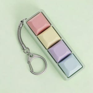 May include: A pastel-colored keychain featuring four square, raised buttons in pink, yellow, purple, and blue. The buttons are set in a light green rectangular frame, attached to a silver spring coil and a keyring.