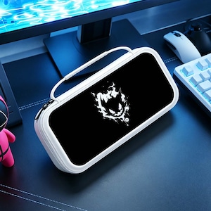 Op de afbeelding: Witte en zwarte draagtas voor een gameconsole. De tas heeft een zwarte voorkant met een wit grafisch ontwerp. De tas heeft een witte rits en een witte handgreep. De tas ligt op een zwart oppervlak.