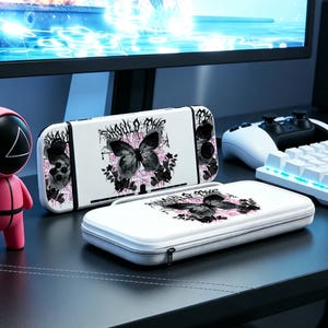 May include: White Nintendo Switch console and carrying case featuring a black and gray butterfly design with pink accents. The console and case are on a black surface, with a computer monitor, controller, and keyboard in the background.
