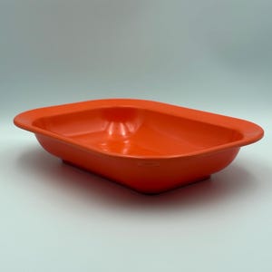 May include: A rectangular baking dish with rounded corners and a smooth, vibrant orange finish. The dish features a wide, flat rim extending outward from the deep, open basin, providing a simple, functional design for food preparation and serving.