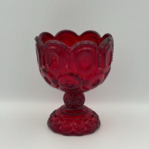 May include: A ruby red glass pedestal bowl. The bowl has a scalloped rim and is decorated with a star and moon pattern. The bowl sits on a pedestal base with a similar star and moon design. The bowl is likely vintage.