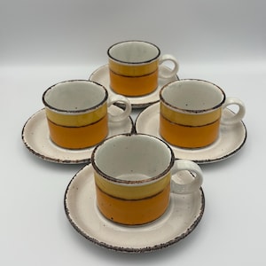 May include: Set of four ceramic espresso cups and saucers. Each cup features a yellow and orange band design with a white interior and a small handle. The saucers are off-white with a speckled texture and a brown rim.