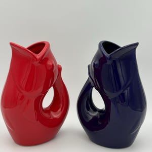 May include: Two ceramic vases, one bright red and the other deep blue. Each vase has a unique, organic shape with a curved handle and a flared opening. The glossy finish enhances the vibrant colors and sculptural design.