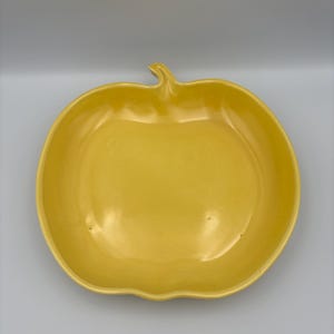 May include: A yellow ceramic serving dish in the shape of an apple. The dish has a stem and a smooth, glossy finish. The dish is a bright, cheerful yellow color.