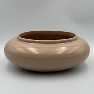 May include: A low, wide, circular ceramic bowl with a smooth, glossy, light beige finish. The vessel features a rounded, bulbous body that tapers inward toward a circular opening at the top, creating a simple, uniform, and continuous silhouette.