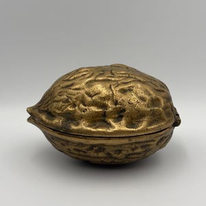 May include: A brass trinket box shaped like a whole walnut. The object features a textured, uneven surface mimicking the natural shell of a walnut, with a horizontal seam running around the middle where the two halves meet and a small hinge.
