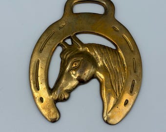 May include: A vintage brass horse head and horseshoe-shaped ornament. The horse head is in profile, centered within the horseshoe. The ornament has a loop at the top for hanging. The brass has a warm, aged patina.