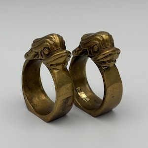 May include: Two brass napkin rings, each featuring a detailed duck head design. The rings have a vintage, aged appearance with a warm, golden hue. The duck heads are intricately sculpted, adding a touch of whimsy to the table setting.