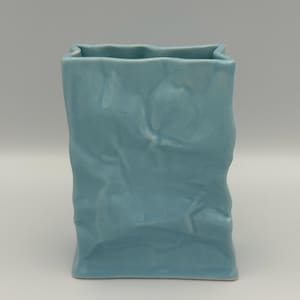 May include: A light blue ceramic vase with a unique crumpled paper bag design. The vase has a rectangular shape with a slightly uneven top edge and textured surface. The color is a soft, pastel blue.