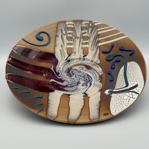 May include: A decorative ceramic plate with a unique abstract design. The plate features a swirl of white and purple in the center, surrounded by brown, burgundy, and beige sections. Accents include blue and white lines and shapes, creating a textured, artistic appearance.