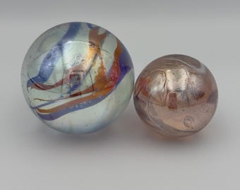 May include: Two glass marbles. One is clear with blue, orange, and purple swirls. The other is a smaller, opaque marble with a pinkish-brown hue and a swirled pattern. Both marbles have a reflective surface.