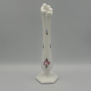 May include: A tall, white, vintage vase with a hexagonal base and a ruffled top. The vase is adorned with delicate pink rose and blue floral accents. The vase is approximately 10 inches tall.