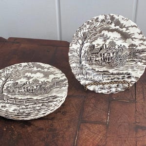 May include: Set of vintage brown and white decorative plates. The plates feature a detailed transferware design of a horse-drawn carriage in a rural landscape. One plate is displayed on a metal stand, while a stack of plates is visible.