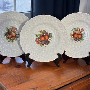 May include: Three white decorative plates with a textured pattern and fruit centerpieces. The plates are displayed on black stands, showcasing a vintage aesthetic. The fruit illustrations feature pears, grapes, and apples.