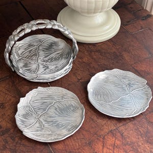 May include: Set of silver-colored coasters with a leaf design, displayed with a matching holder featuring a floral handle. The coasters are round and have a textured surface. A white decorative vase is in the background.