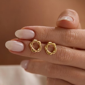 May include: A pair of small, gold-colored stud earrings. Each earring features a woven, circular design. The earrings are displayed on a person's hand, showcasing their delicate and intricate craftsmanship. The earrings are a simple yet elegant accessory.
