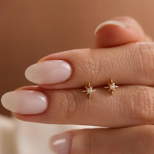 May include: A pair of small, gold-colored starburst stud earrings. Each earring features a central, clear gemstone. The earrings are held between the fingers of a person.