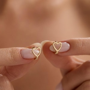 Può includere: Un paio di orecchini a cerchio a forma di cuore color oro. Gli orecchini sono tenuti tra le dita, mettendo in risalto il loro design delicato. La forma a cuore è aperta, creando un'estetica minimalista ed elegante. Gli orecchini sono un classico della gioielleria.