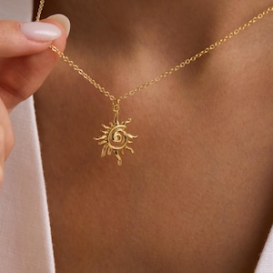 Pode incluir: Um colar dourado com um pingente de sol. O pingente de sol tem um centro em espiral e raios que se estendem para fora. O colar está sendo segurado por uma pessoa. A corrente é delicada e o pingente é pequeno.