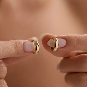 Può includere: Un paio di piccoli orecchini a cerchio dorati. Gli orecchini sono tenuti tra due mani con unghie curate. Gli orecchini hanno un design semplice e arrotondato e una finitura lucida. Lo sfondo è sfocato.