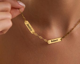 May include: A gold-colored necklace with two rectangular nameplates. One plate reads "Jessica" and the other reads "Robert". The necklace has a delicate chain design. The jewelry is displayed against a neutral background.