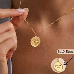 May include: A gold necklace featuring a round pendant with a compass design. The pendant hangs from a delicate chain. The image shows a close-up of the pendant's back, which can be engraved. The text "Back Engraving" is also visible.