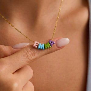 May include: A gold chain necklace spelling out the name "EMMA" in colourful, block-style letters. The letters are pink, blue, green, and purple. The necklace is held up by a finger.