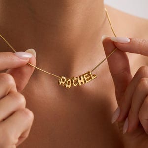 May include: A gold-colored necklace with the name "RACHEL" spelled out in individual, block-style letters. The necklace is held by a person, showcasing the delicate chain and the personalized pendant. The background is a soft, neutral tone.