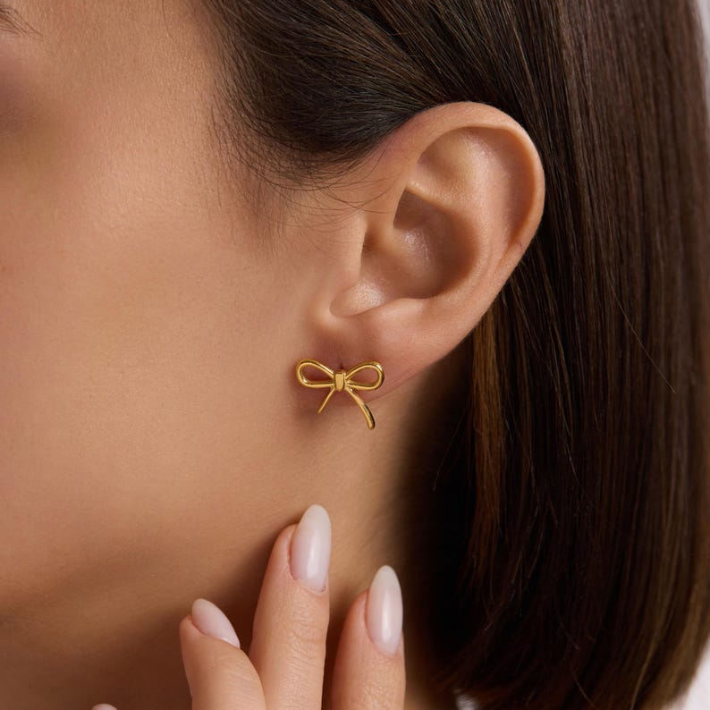 May include: A pair of gold-coloured bow-shaped stud earrings. The earrings are small and delicate, with a simple, elegant design. The bows are symmetrical and have a polished finish. The earrings are shown on an ear.