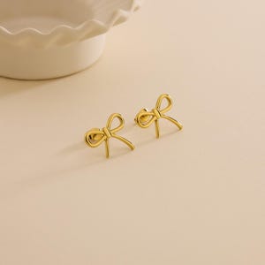 May include: A pair of gold-coloured bow-shaped stud earrings. The earrings are small and delicate, with a simple, elegant design. They are displayed on a light-coloured surface, with a white, scalloped-edge dish in the background.