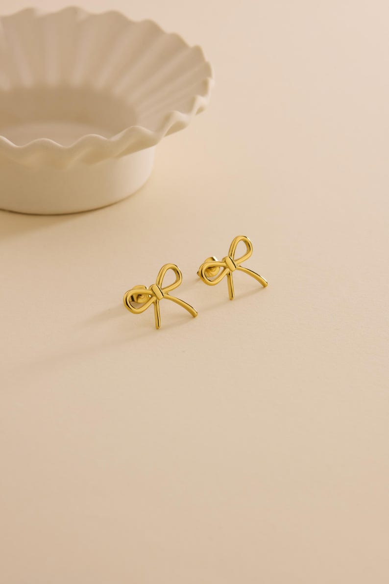 May include: A pair of gold-coloured bow-shaped stud earrings. The earrings are small and delicate, with a simple, elegant design. They are displayed on a light-coloured surface, with a white, scalloped-edge dish in the background.