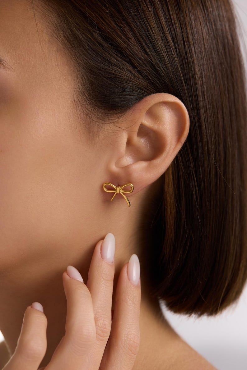 May include: A pair of gold-coloured bow-shaped stud earrings. The earrings are small and delicate, with a simple, elegant design. The bow shape is clearly defined, with the loops and tails of the bow visible. The earrings are shown on an ear.
