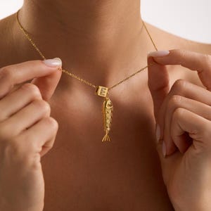 Könnte beinhalten: Goldfarbene Halskette mit einem Fischanhänger und einem Würfel mit dem Buchstaben "E". Der Fischanhänger hat detaillierte Schuppen und Flossen. Die Halskette wird von einer Person gehalten.