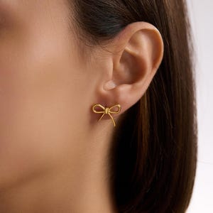 May include: A close-up of an ear with a gold bow-shaped earring. The earring is a small, delicate design, with a bow shape. The earring is made of a shiny gold material.
