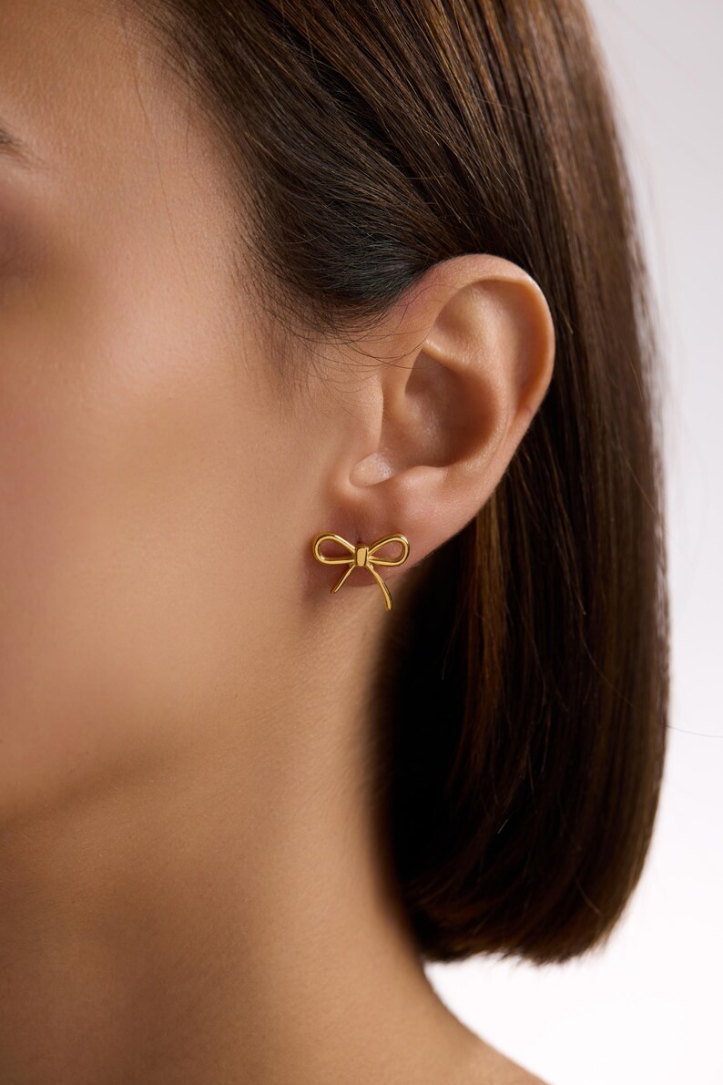 May include: A close-up of an ear with a gold bow-shaped earring. The earring is a small, delicate design, with a bow shape. The earring is made of a shiny gold material.