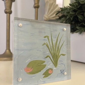 May include: Square acrylic painting featuring a water lily scene. The artwork depicts green lily pads, pink flowers, and tall green reeds against a light blue background. The painting is secured with four small, white fasteners.