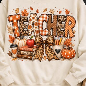 Op de afbeelding: Een crèmekleurige sweater met ronde hals en een grote afbeelding met het woord TEACHER in diverse patronen. Het ontwerp bevat herfstbladeren, een stapel boeken, een pompoen, een koffiebeker, een pennenhouder en een strik met luipaardprint.