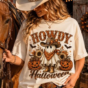 Pode incluir: Uma t-shirt de cor creme com um gráfico de um fantasma a usar um chapéu de cowboy e bandana, rodeado por girassóis e abóboras. O texto "HOWDY" e "Halloween" está impresso em laranja e com um padrão de leopardo.
