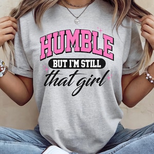 May include: A light gray t-shirt with the text "HUMBLE" in pink block letters, outlined in black, above the words "BUT I'M STILL" in white on a black oval, and "that girl" in cursive. The shirt is a casual style.