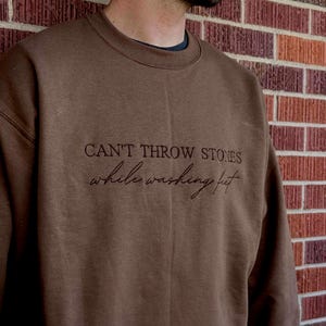 May include: A brown sweatshirt with the embroidered text "CAN'T THROW STONES while washing feet." The crew neck sweatshirt is a solid color and has a relaxed fit. The background is a brick wall.